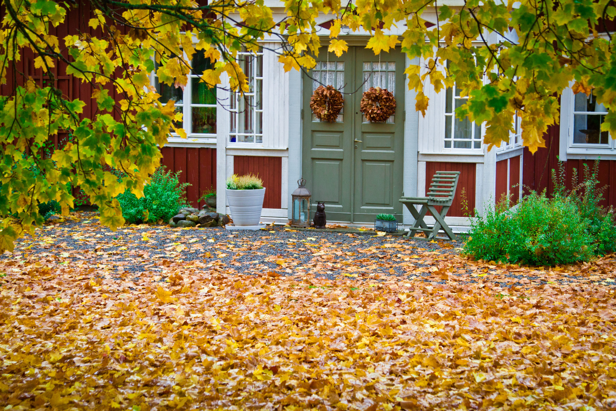 Ett rött trähus med en grön dubbeldörr. På dörrfönstren hänger två lövkransar. Bredvid dörren står det en grön trästol. På marken ligger det fullt med gula lönnlöv.