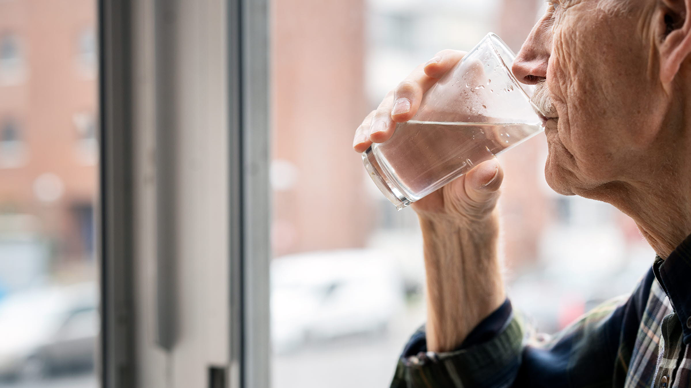 Äldra person i profil som dricker ur ett vattenglas och ser ut genom ett fönster